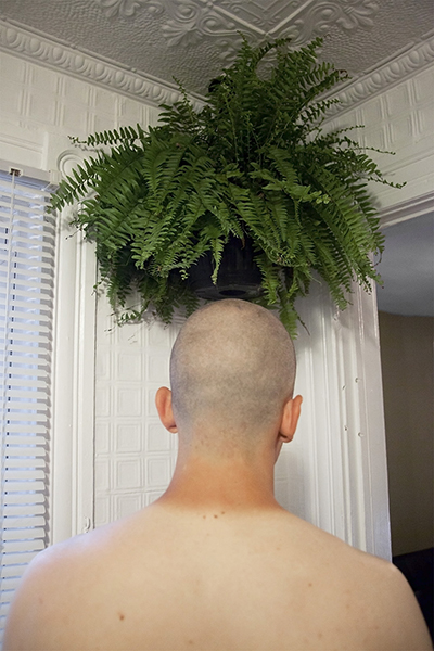 20" x 30" Archival Inkjet Print. Color photograph. Back of a young man's head, shaved, tan line, fern, plant, corner of a room